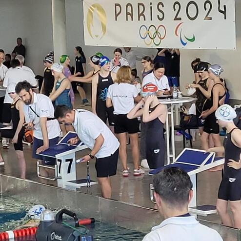 Wettkampfszenario mit Schwimmern am Beckenrand, Startblöcken und einem Banner für die Olympischen Spiele Paris 2024.