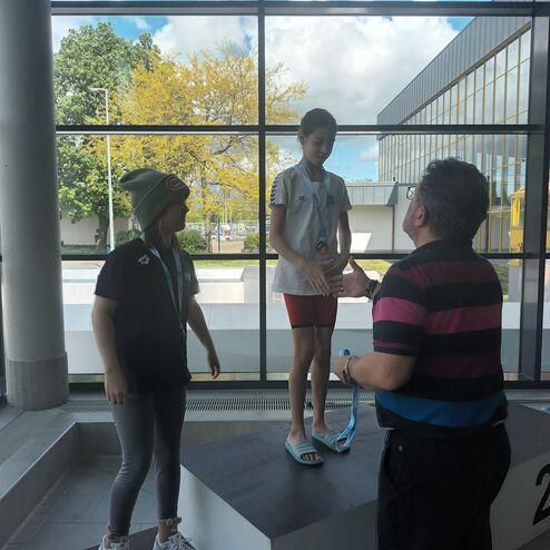 Zwei Kinder auf einem Siegerpodest; eines erhält eine Medaille von einem Erwachsenen, im Hintergrund große Fenster.