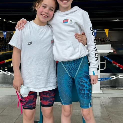 Zwei Mädchen stehen lächelnd in Freizeitkleidung am Schwimmbeckenrand, umarmt und bereit für den Wettbewerb.