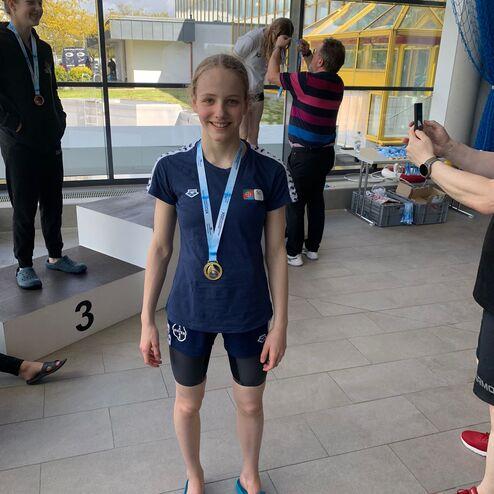 Ein Mädchen in Schwimmbekleidung mit Medaille lächelt vor einer Glaswand, während andere Personen im Hintergrund jubeln.
