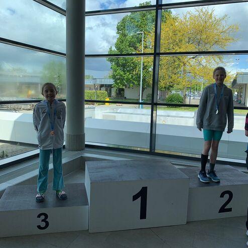 Zwei Mädchen mit Medaillen stehen auf einem Siegertreppchen, umgeben von großen Fenstern und grünem Laub.