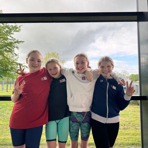 Vier Mädchen posieren lächelnd vor einem Fenster, tragen Schwimmbekleidung und zeigen Friedenzeichen mit den Händen.