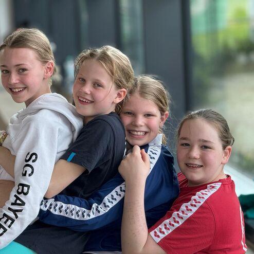 Vier Mädchen lächeln und stehen eng beieinander, sie tragen Sportbekleidung und sitzen auf einer Bank.
