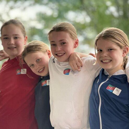 Vier junge Mädchen stehen nebeneinander und lächeln, während sie sich freundschaftlich umarmen, mit Namensschildern auf ihren Sportanzügen.