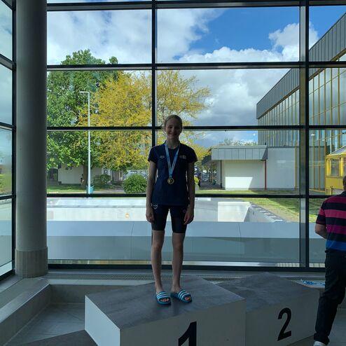 Mädchen steht auf dem ersten Podest mit Medaille, vor großen Fenstern und sichtbarem Schwimmbecken im Hintergrund.