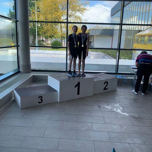 Zwei Kinder mit Medaillen stehen auf dem Siegertreppchen, mit Platz eins, zwei und drei sichtbar davor.