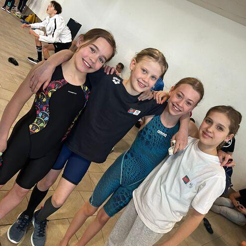 Vier Mädchen posieren lächelnd in Badesportbekleidung, umarmt im Innenbereich eines Schwimmbads.