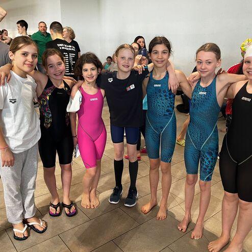 Eine Gruppe von sechs Mädchen in Schwimmanzügen posiert lächelnd für ein Foto in einem Schwimmveranstaltungsraum.