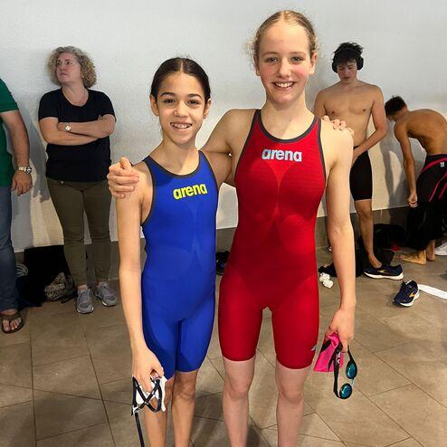 Zwei junge Schwimmerinnen posieren in Badeanzügen, eine in Blau und eine in Rot, mit Schwimmbrillen in der Hand.