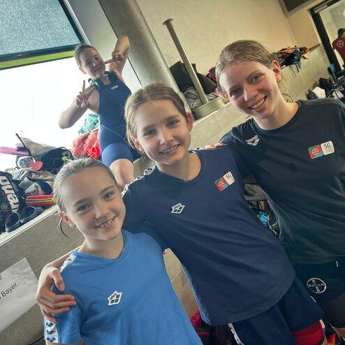 Drei Mädchen in Schwimmkleidung lächeln gemeinsam vor einem Tisch mit Taschen im Hintergrund.