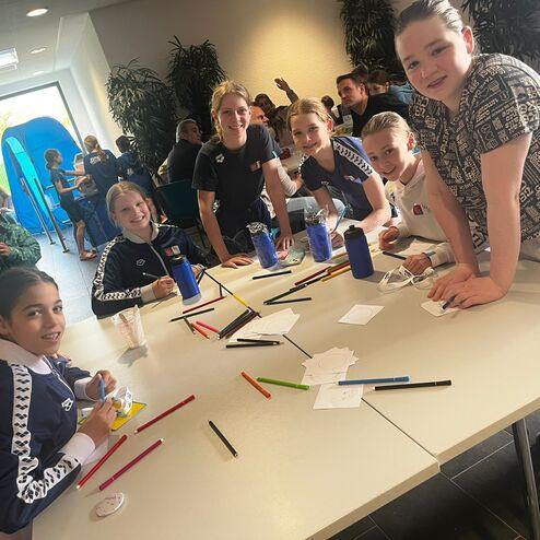 Gruppe von Kindern bei kreativer Tätigkeit am Tisch, umgeben von Stiften und Zeichenmaterial in einem Raum.