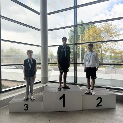Drei Jugendliche stehen auf einem Siegerpodest mit Medaillen und einem modernen Fensterhintergrund.