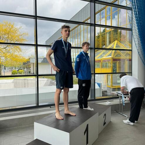 Zwei Sportler stehen auf einem Siegertreppchen, der eine mit Medaille, im Hintergrund große Fenster und gelbe Bäume.