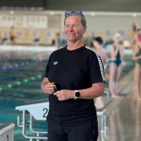 Trainerin mit Stoppuhr an einem Schwimmbecken, im Hintergrund schwimmende Athleten und Startblöcke.
