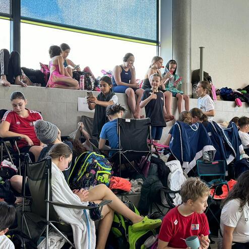 Gruppe von Kindern in Badebekleidung und T-Shirts, die auf Stühlen sitzen und entspannen, im Hintergrund eine große Fensterfront.