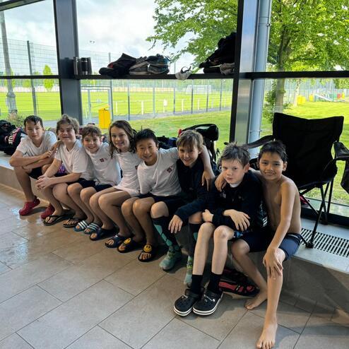 Gruppe von acht Kindern in Poolkleidung sitzen an einer Fenstermauer, Blick auf Sportplatz im Hintergrund.