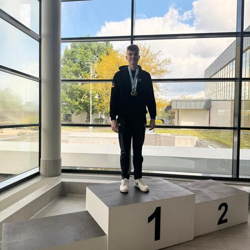 Junge steht auf dem ersten Platz eines Siegertreppchens und trägt drei Medaillen, sichtbar durch ein großes Fenster.
