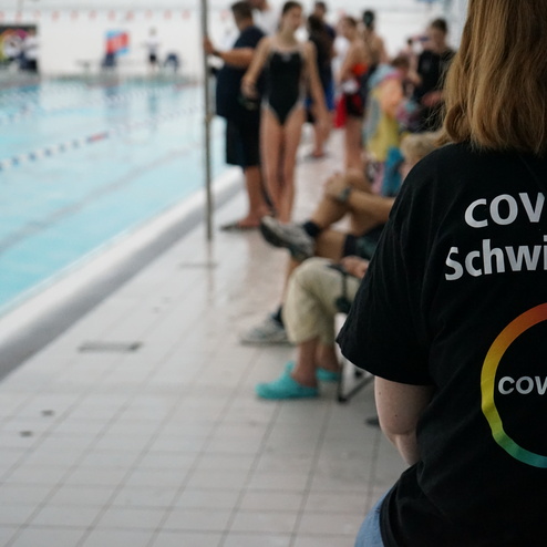 Person mit T-Shirt "Covestro Schwimmfest" steht neben einem Schwimmbecken, im Hintergrund Zuschauer und Schwimmer.