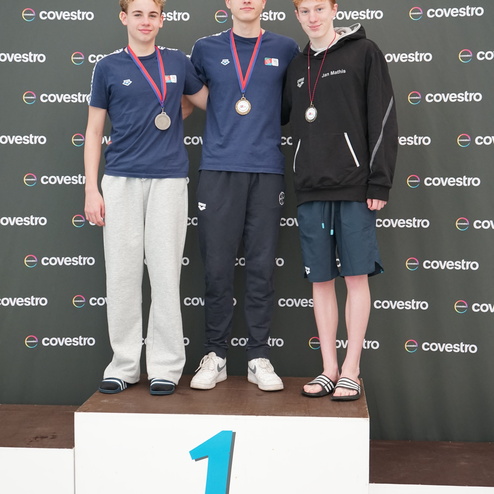 Drei Medaillengewinner stehen auf einem Siegerpodest mit der Nummer 1, im Hintergrund ein Covestro Banner.