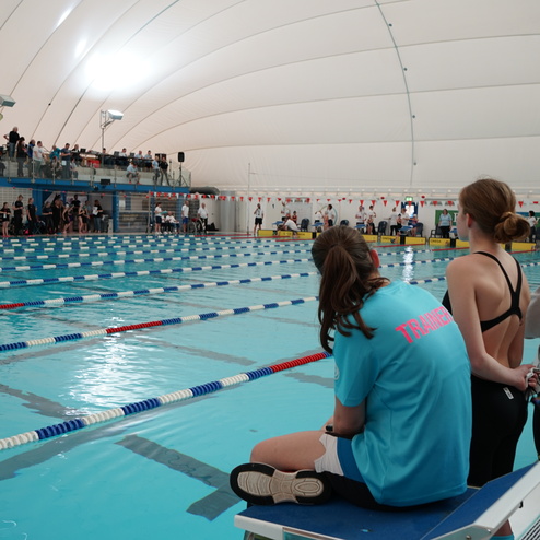 Schwimmhalle mit Schwimmern und Zuschauern; Trainerin sitzt auf einem Podest am Beckenrand und beobachtet das Geschehen.