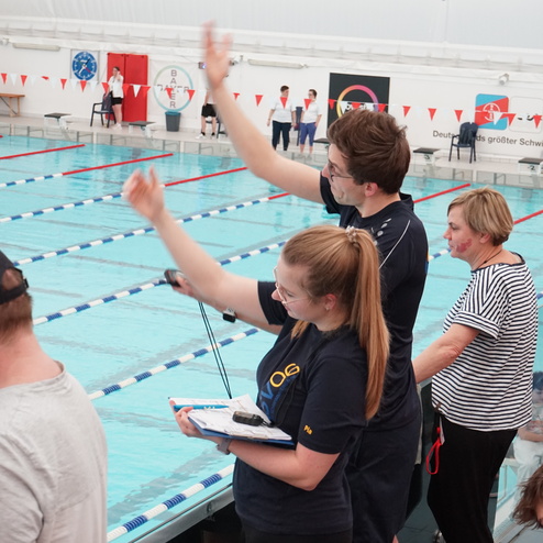 Zwei Aufsichtspersonen heben die Hand am Beckenrand eines Schwimmbeckens, während Zuschauer im Hintergrund stehen.