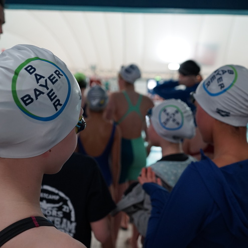 Gruppe von Schwimmern in Badekappen mit Logo, die sich auf ein Training am Schwimmbecken vorbeireiten.