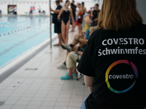 Person mit T-Shirt "Covestro Schwimmfest" steht neben einem Schwimmbecken, im Hintergrund Zuschauer und Schwimmer.