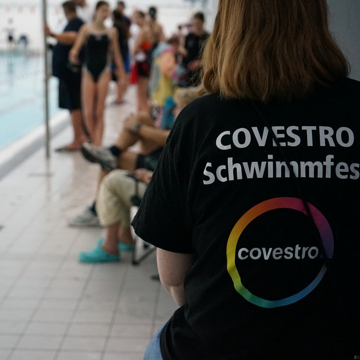 Person mit T-Shirt "Covestro Schwimmfest" steht neben einem Schwimmbecken, im Hintergrund Zuschauer und Schwimmer.
