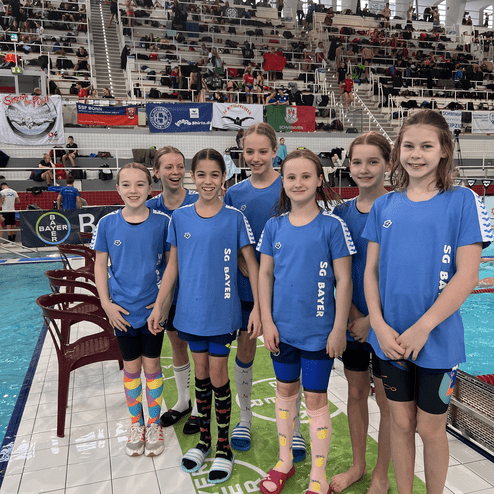 Gruppe von sieben jungen Schwimmerinnen in blauen T-Shirts, stehen am Beckenrand, umgeben von Zuschauern und Bannern.