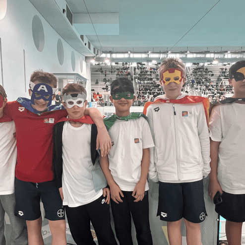 Sechs Kinder in Superheldenmasken und Umhängen stehen fröhlich neben einem Schwimmbecken in einem Stadion.