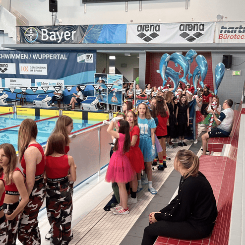 Gruppe von Kindern in bunten Kostümen wartet an einem Schwimmbecken in einer Sporthalle, einige halten Luftballons.