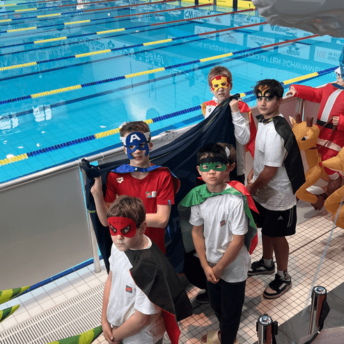 Gruppe von fünf Kindern in Superheldenkostümen und Masken vor einem Schwimmbecken, bereit für ein Wettkampf-Event.
