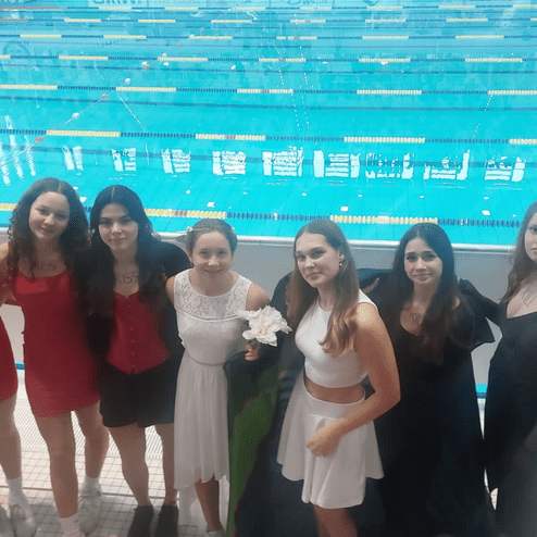 Gruppe von acht jungen Frauen in festlicher Kleidung vor einem Schwimmbecken mit blauen Wasser und Startblöcken.
