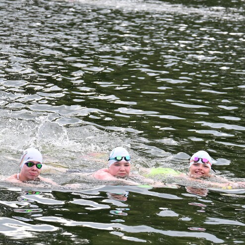 Drei Schwimmer in Badekappen und Sonnenbrillen gleiten durch grünes Wasser, umgeben von sanften Wellen.