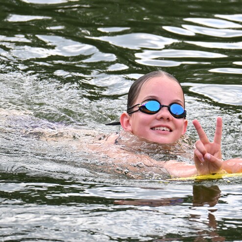 Ein Mädchen schwimmt im Wasser und zeigt ein Siegzeichen, trägt Schwimmbrille und hält ein Schwimmbrett.