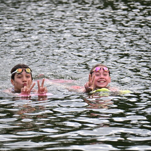 Zwei Kinder im Wasser zeigen das Peace-Zeichen und lächeln, während sie in einer ruhigen Gewässeroberfläche schwimmen.