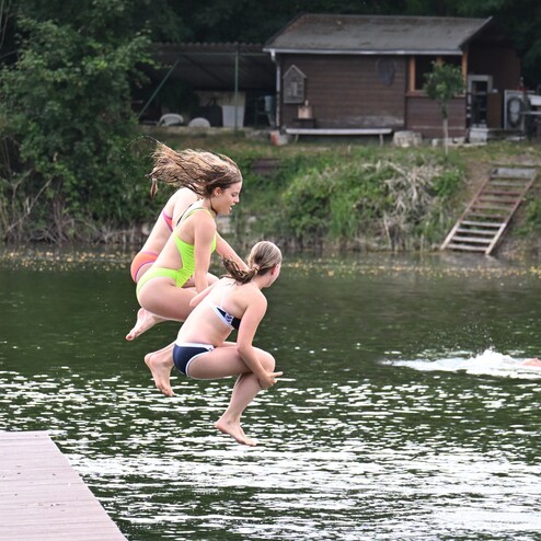 Zwei Mädchen springen synchron ins Wasser von einem Holzsteg an einem ruhigen Gewässer.