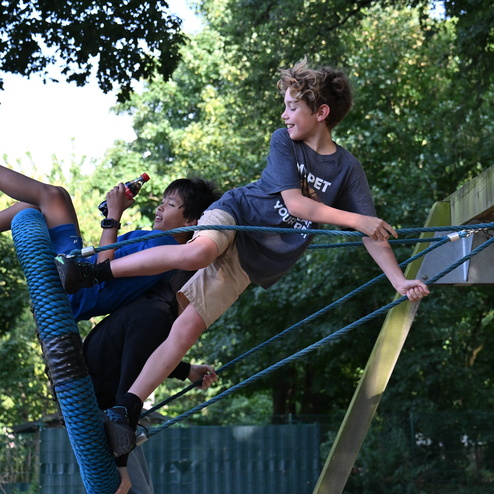 Zwei Kinder sitzen auf einem Seilspielzeug, eines hält eine Flasche. Im Hintergrund sind Bäume sichtbar.