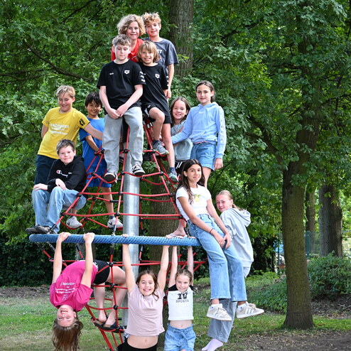 Gruppe von Kindern klettert auf einem Spielturm aus Seilen und posiert in einem Park mit Bäumen im Hintergrund.