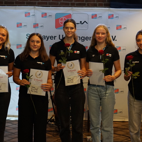 Fünf weibliche Sportlerinnen halten Urkunden und rote Rosen bei einer Verleihung in einem Veranstaltungsraum.