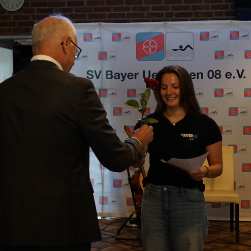 Ein älterer Mann überreicht einer jungen Frau mit Rose in der Hand ein Zertifikat auf einer Veranstaltung.