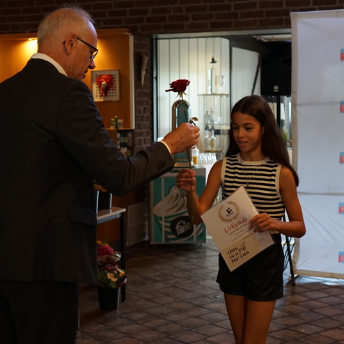 Ein älterer Mann überreicht einem Mädchen eine rote Rose und eine Urkunde in einem festlichen Raum.
