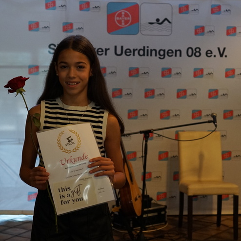 Mädchen mit braunen Haaren hält eine Urkunde und eine Rose vor einem Veranstaltungsbanner.