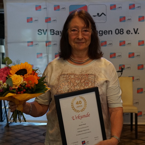 Frau hält einen bunten Blumenstrauß und eine Urkunde für 40 Jahre Vereinsmitgliedschaft in einem festlichen Raum.