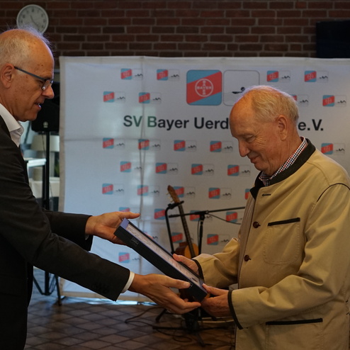 Zwei Männer überreichen sich eine Urkunde während einer Veranstaltung, im Hintergrund ein Banner des SV Bayer Uerdingen.