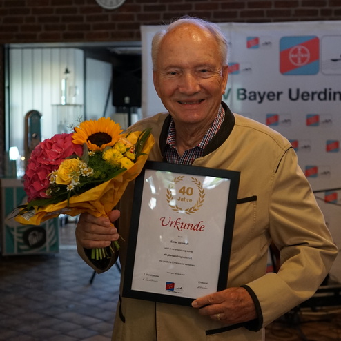 Ein älterer Mann hält eine Urkunde für 40 Jahre Mitgliedschaft und einen Blumenstrauß in einer festlichen Umgebung.