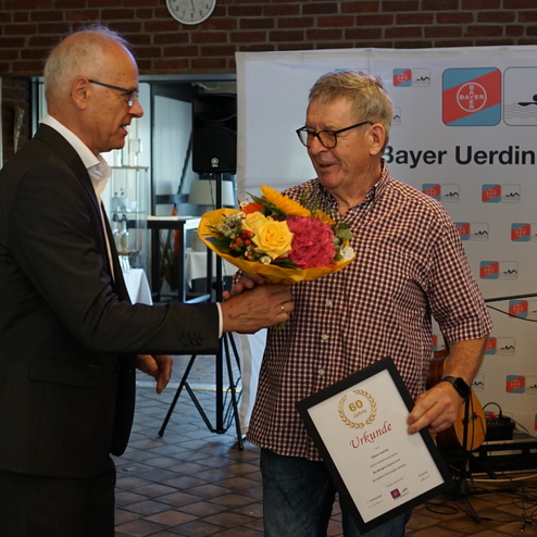 Zwei Männer in einem Festsaal überreichen ein Blumenbouquet und eine Urkunde zum 60. Jubiläum eines Vereins.