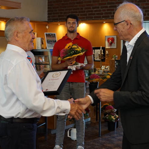 Zwei Männer schütteln sich die Hände, während einer einen Blumenstrauß hält; im Hintergrund eine feste Umgebung mit Dekoration.