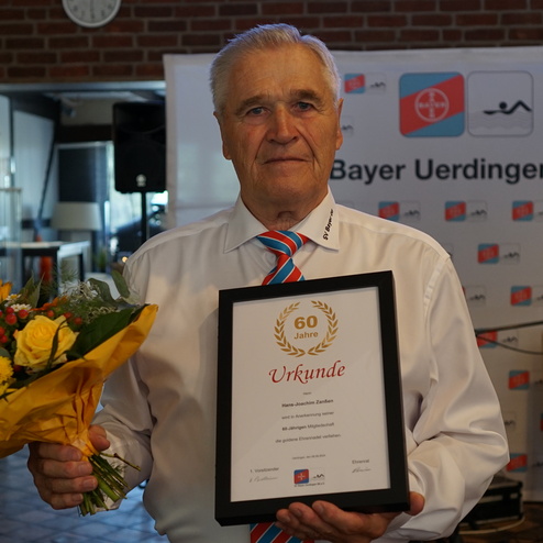 Älterer Mann in weißem Hemd hält eine Urkunde und einen Blumenstrauß bei einer Ehrenveranstaltung.