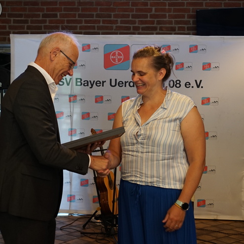 Ein Mann und eine Frau schütteln sich die Hände bei einer Auszeichnung in einem festlichen Raum mit Hintergrundbanner.